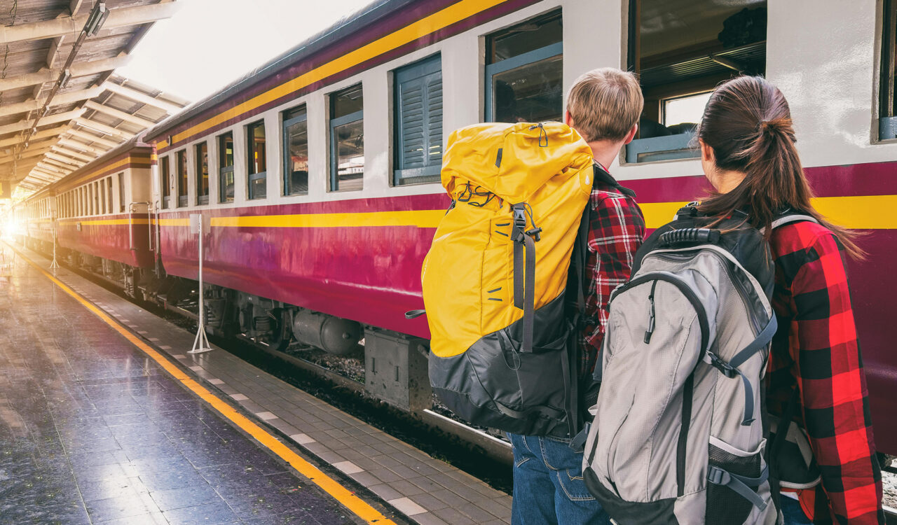 2 Jugendliche mit Rucksack von hinten fotografiert, die vor einem Zug am Bahnhof stehen.