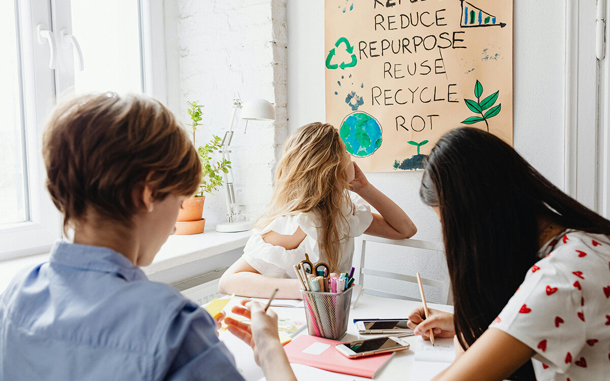 Junge Frauen sitzen um einen Tisch herum und schreiben, eine dreht sich zu einem Plakat dahinter um, auf dem englische Begriffe zu Umweltschutz stehen.