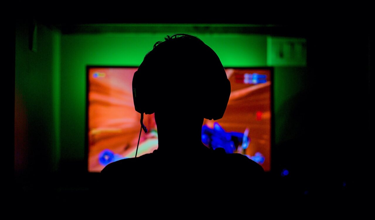 Silhouette eines jungen Mannes, der Kopfhörer auf hat und vor einem Screen sitzt und Computer spielt.