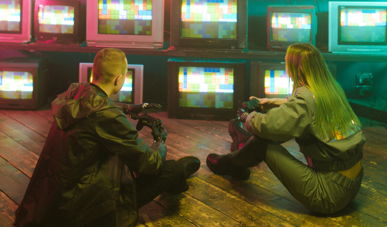 Zwei Jugendliche sitzen am Boden vor einer Wand mit vielen Monitoren.