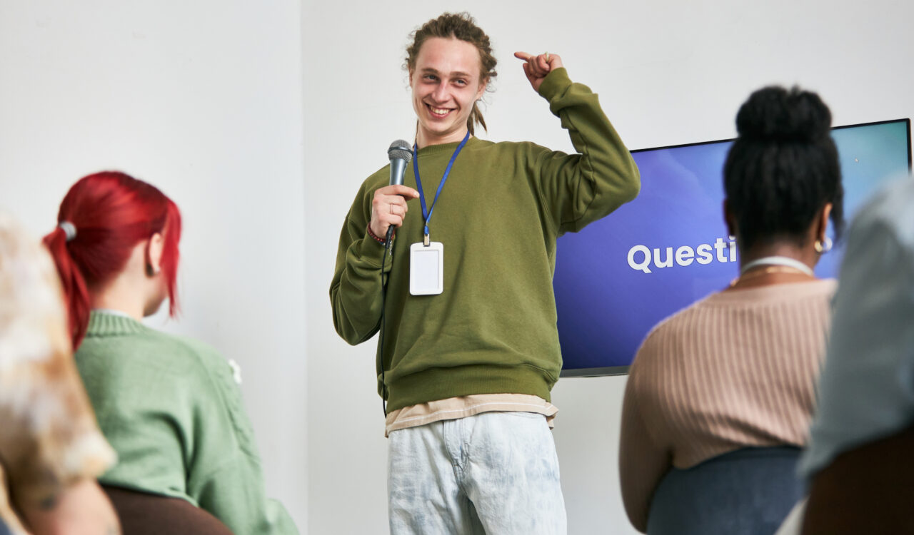 Junger Mann steht mit Mikrofon da, lächelt und interagiert während einer Präsentation mit einem vielfältigen Publikum.