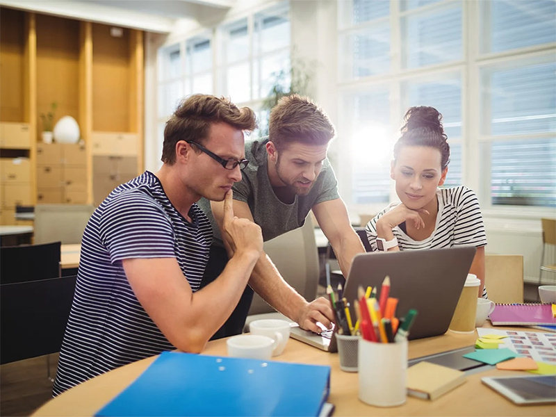 2 Männer und eine Frau schauen auf einen Laptop, der vor ihnen am Schreibtisch steht.
