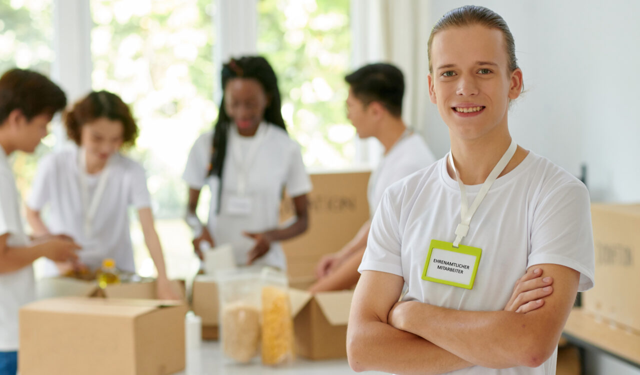 Porträt eines lächelnden Freiwilligen einer Wohltätigkeitsorganisation, sein Freund verpackt gespendete Produkte im Hintergrund.