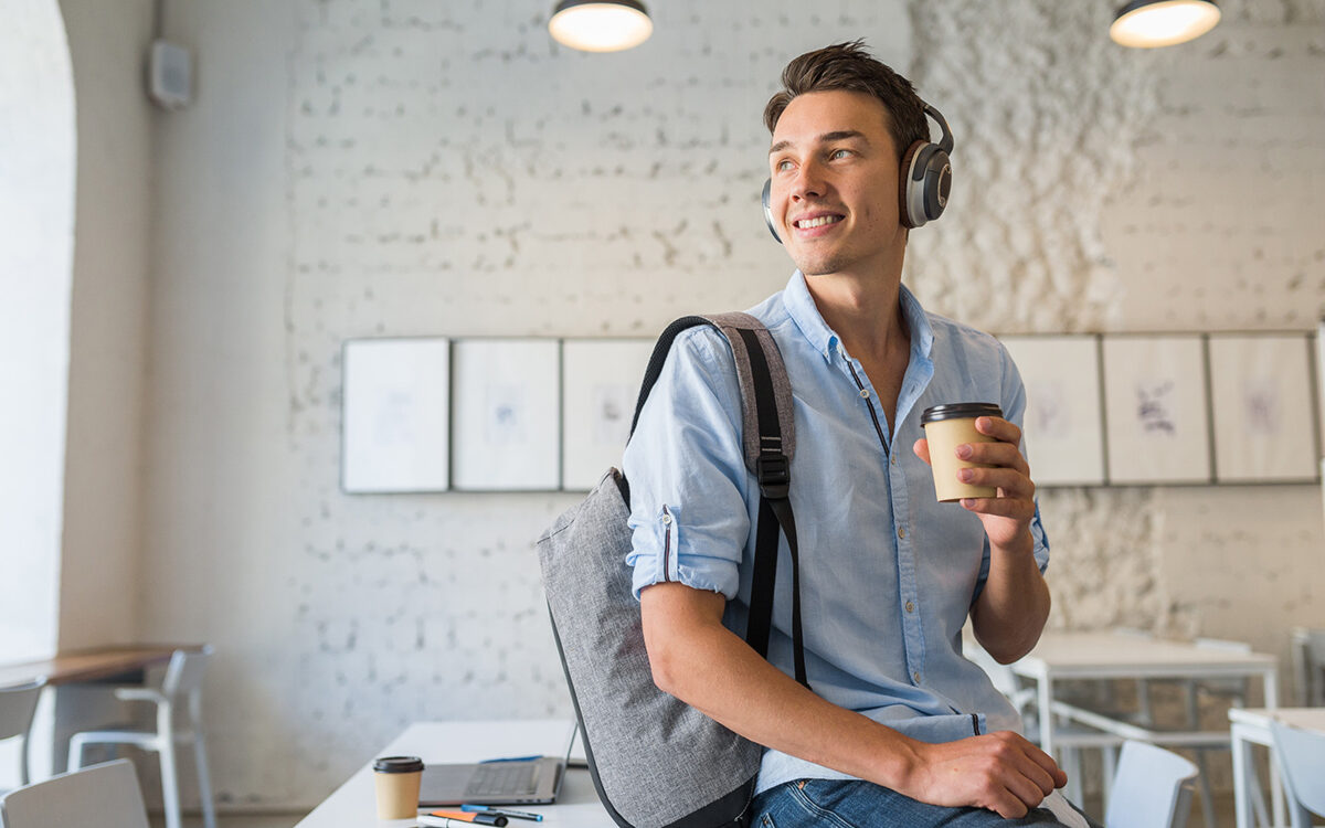 Junger Mann sitzt auf einem Tisch in einer Agentur, er hat Kopfhörer auf und einen Kaffeebecher in der Hand.