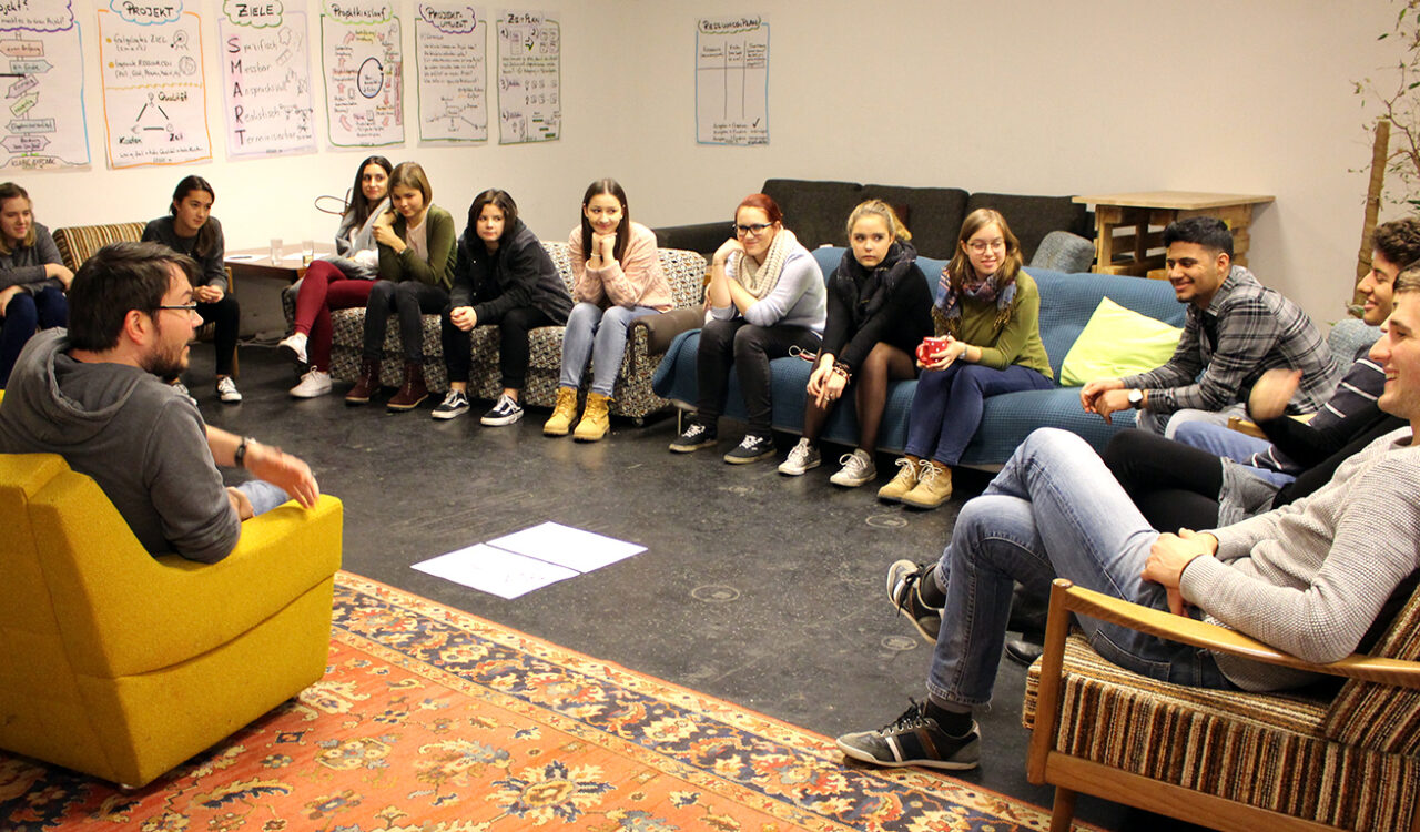 Eine Gruppe jugendlicher Personen sitzen in einem Sesselkreis und diskutieren.