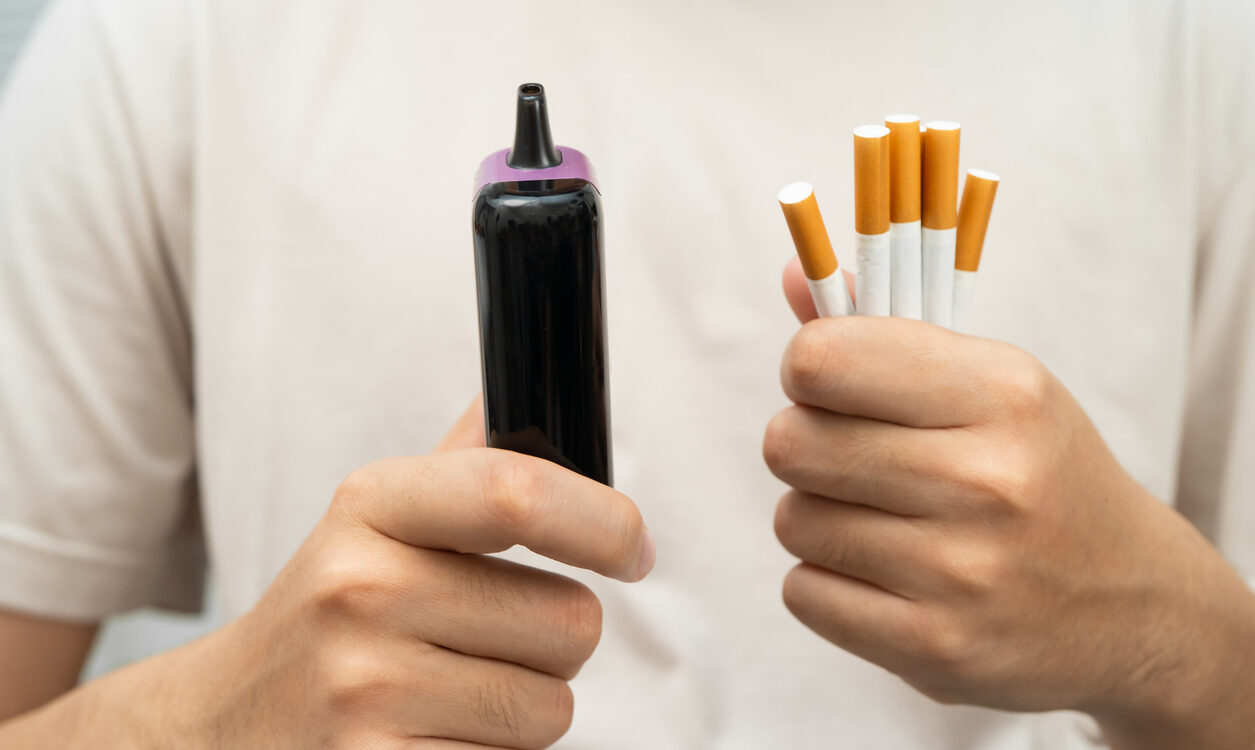 Ein junger Mann hält in einer Hand mehrere Zigaretten, in der anderen Hand eine elektronische Zigarette oder Vape.