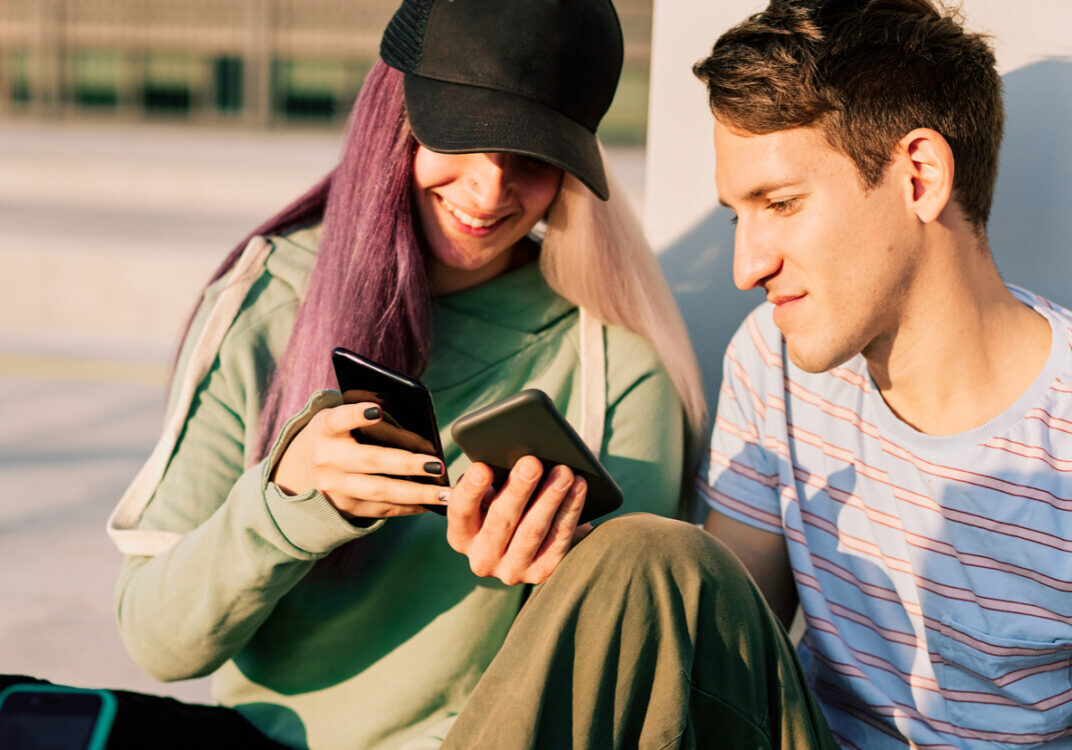 Eine kleine Gruppe von jugendlichen Personen aller Geschlechter sitzen im Freien und schauen zusammen auf Mobiltelefone (Handys). Die jugendlichen Personen sind gut gelaunt.